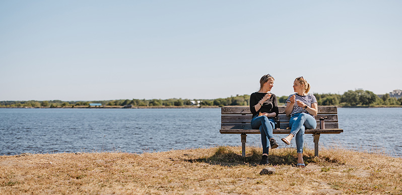 Två kvinnor sitter på en träbänk med havet som bakgrund.